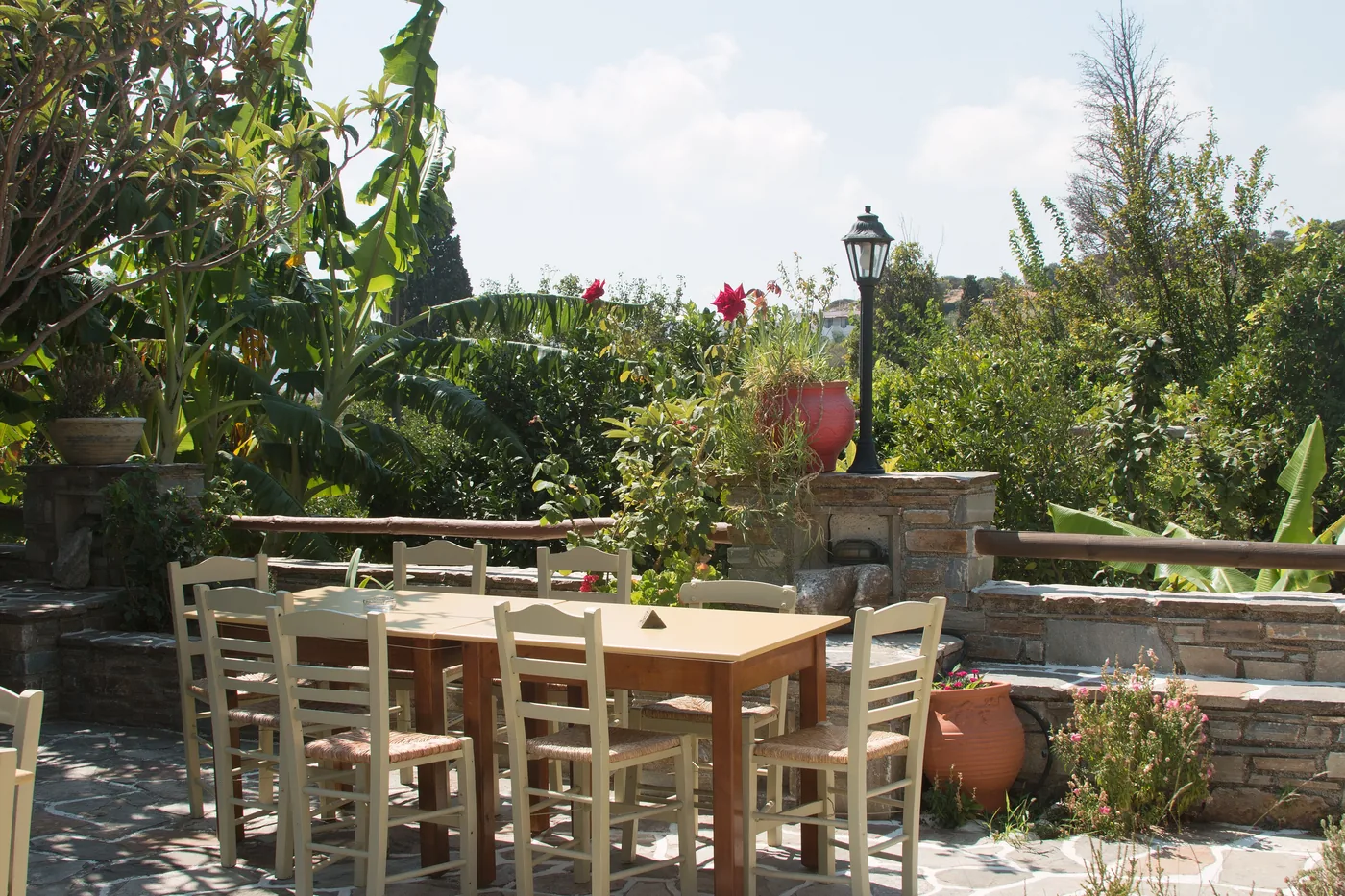 A shaded stone terrace in Ano Potamia with a wooden table, wicker-seated chairs, potted geraniums and a lush garden behind a low wall